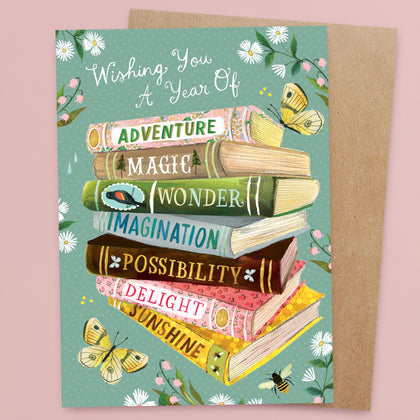 Illustrated card with stacked books labeled Adventure, Magic, Wonder, Imagination, Possibility, Delight, and Sunshine. Text above reads Wishing You A Year Of. Butterflies, flowers, and a bee decorate the design.