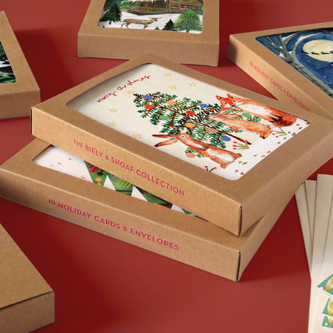 Stacked boxes of holiday cards featuring festive illustrations, including a Christmas tree with deer, placed on a red surface. Other card boxes and envelopes are visible nearby.
