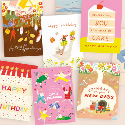 A woman in a floral dress sits at a table with greeting cards, pens, a mug, and a vase of pink tulips, smiling at the camera. Art supplies and colorful cards are on the wall behind her.