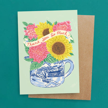 A greeting card with an illustration of a fluffy sheep holding a colorful bouquet of flowers in its mouth. The text above the sheep reads, thank ewe.