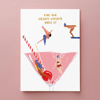Illustration of four women in swimsuits diving and swimming in a giant pink cocktail glass with a cherry, straw, and lemon; text above reads, MAKE YOUR BIRTHDAY HANGOVER WORTH IT!.