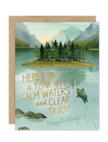 The Clear Skies Birthday Card by Biely & Shoaf features a serene lake, mountains, and a canoeist paddling toward a wooded island. Printed on recycled paper, it reads: Here’s to a year of calm waters and clear skies! Happy Birthday!.