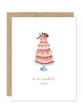 The Wedding Cake Luxe Wedding Card by Biely & Shoaf features a gold foil three-tiered cake with flowers on a blue stand and “to the beautiful couple” in gold cursive. Part of the A2 Luxe Collection, includes a brown envelope.