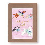 Biely & Shoaf’s Dogs Making Spirits Bright Boxed Holiday Cards feature four festive dogs in sweaters on a pink background with gold stars and foil text. The FSC-certified brown box has a clear window to display the card design.