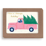 The Tree-rific Penguin In Truck Boxed Holiday Cards by Biely & Shoaf feature a vintage pink truck with a penguin driver and Christmas trees, are packaged in brown recycled paper, and include 10 holiday cards saying “Have a treerific holiday!”.