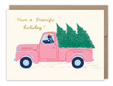 Illustration of a pink pickup truck labeled “Blitzen,” carrying Christmas trees and driven by a penguin in a red hat. Have a treerrific holiday! appears above. Biely & Shoaf’s Tree-rific Penguin In Truck Holiday Card features gold foil accents.