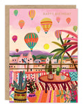 The Hot Air Balloons Birthday Card by Biely & Shoaf features colorful balloons floating above a vibrant town at sunset, with patterned rugs, potted plants, and a festive table on FSC-certified recycled paper.