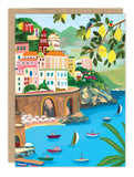 The Amalfi Coast Birthday Card by Biely & Shoaf features vibrant cliffside buildings, sailboats, kayaks on blue water, lush greenery, two birds on a lemon tree branch, and gold foil “Happy Birthday” text. Illustrated by Hebe Studio.