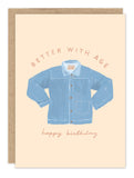 The Biely & Shoaf Better With Age Denim Birthday Card features a Karen Schipper illustration of a blue denim jacket with "BETTER WITH AGE" above and "happy birthday" in cursive below, displayed on a brown envelope.