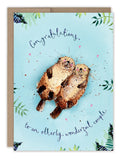 The Biely & Shoaf Otterly Wonderful Couple Anniversary Card features two otters cuddling among green leaves on a blue background, with the message “Congratulations, to an otterly wonderful couple” and pink hearts—perfect for anniversaries.