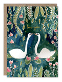 The Swans Love Card by Biely & Shoaf features two white swans facing each other on a dark green floral background with gold "love you" text. It includes a brown envelope and is printed on FSC-certified recycled paper.
