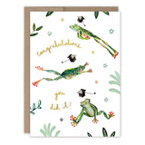 The Frogs Graduation Card by Biely & Shoaf features illustrated frogs jumping among caps and leaves, with “Congratulations you did it!” in handwritten text on white recycled paper. Includes a brown envelope.