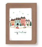 The Little Christmas Village Boxed Holiday Cards by Biely & Shoaf feature a festive scene of colorful townhouses, Christmas trees, people, and a red car in the snow with “Merry Christmas” below. Cards are in a recycled paper brown box.