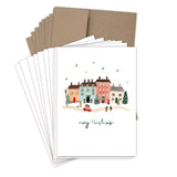 The Little Christmas Village Boxed Holiday Cards by Biely & Shoaf feature festive townhouses, people, a decorated tree, and snowflakes. "Merry Christmas" is written in green cursive; brown envelopes are included.