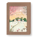 The Biely & Shoaf Red Sky Cozy Christmas Boxed Holiday Cards feature an illustrated snowy village at sunset with houses, trees, people, and a dog. The cards read "warm and cozy Christmas wishes" and come in a brown box printed on recycled paper.
