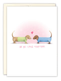 The Be-long Together Hounds Anniversary Card by Biely & Shoaf features two dachshunds in sweaters facing each other with a pink heart, small pink hearts on a white background, and “WE BE-LONG TOGETHER” text—a sweet anniversary greeting.