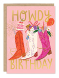 The Biely & Shoaf Howdy Birthday Card features three cowboy boots with birthday motifs (cake, gift, flowers) on a pink, gold-foiled card with "Howdy Birthday" in bold gold lettering above and below the boots.
